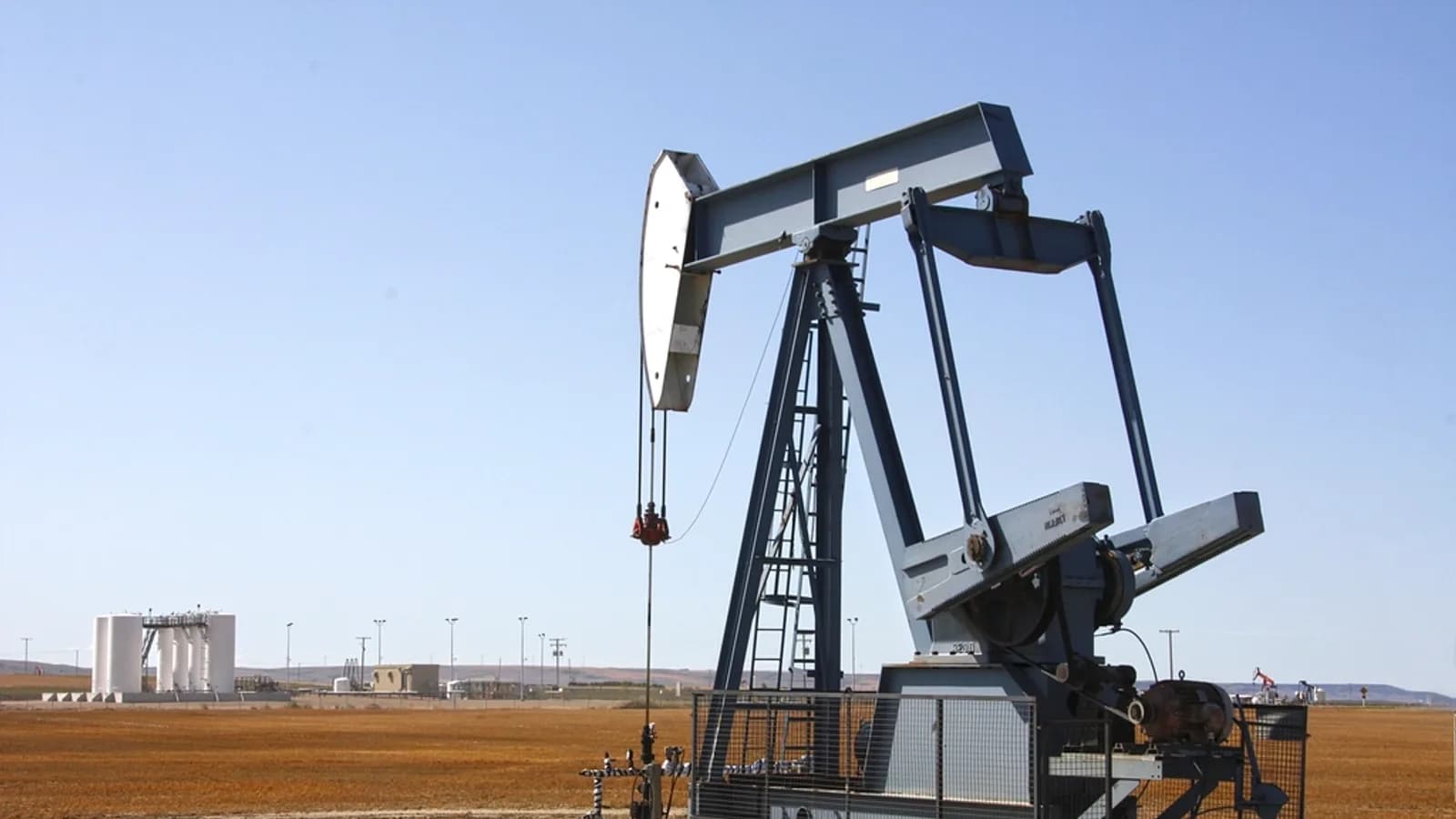 A pumpjack on an oil lease in flat open country, with a processing facility on the horizon