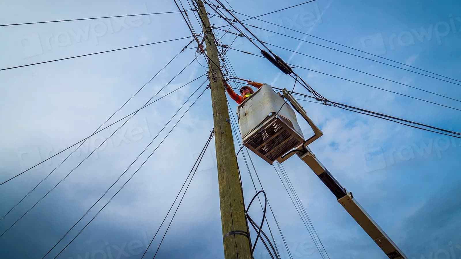 A technician working from a bucket at the top of a utility pole among cables