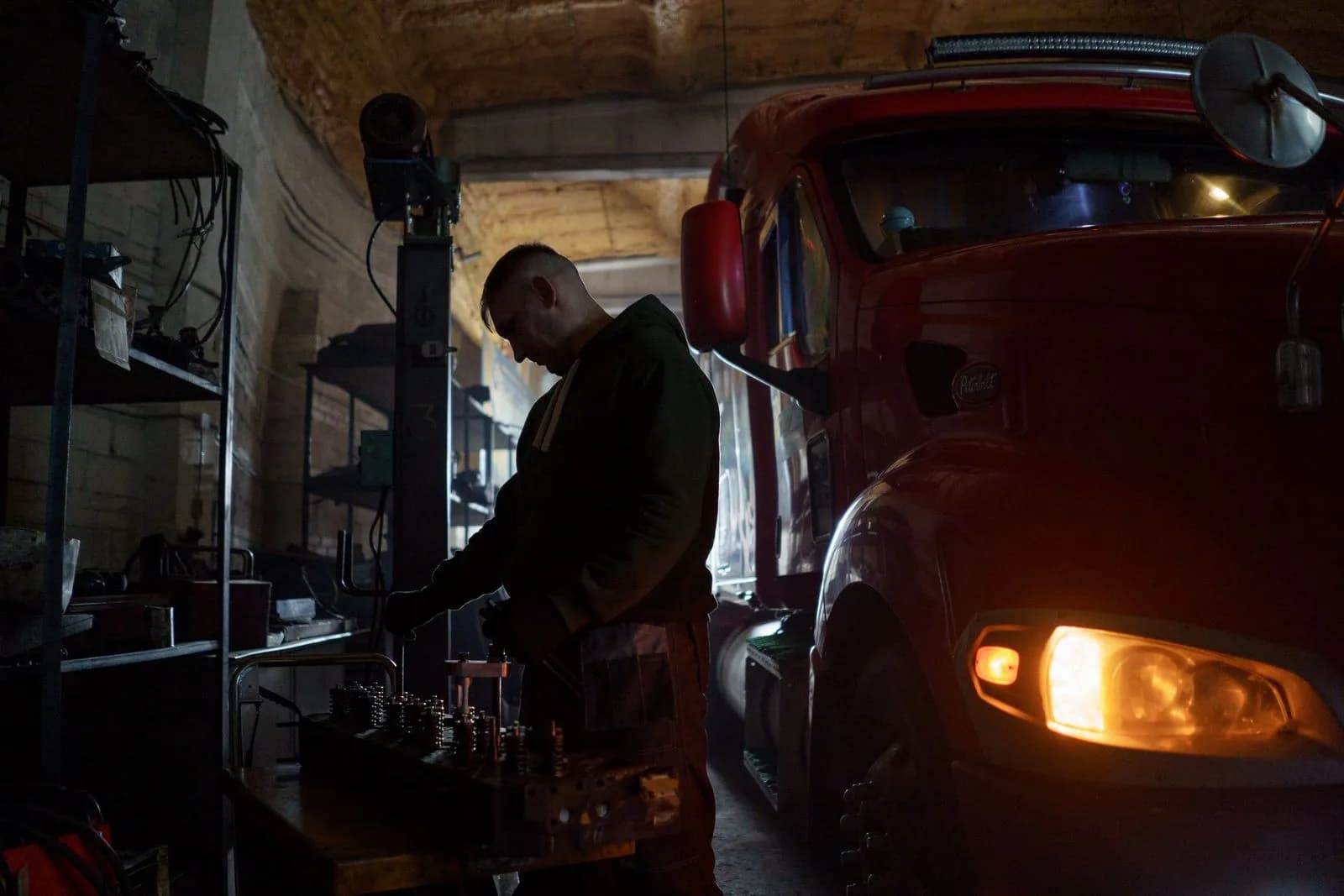 A mechanic working at a bench beside a red semi-truck in a dim garage