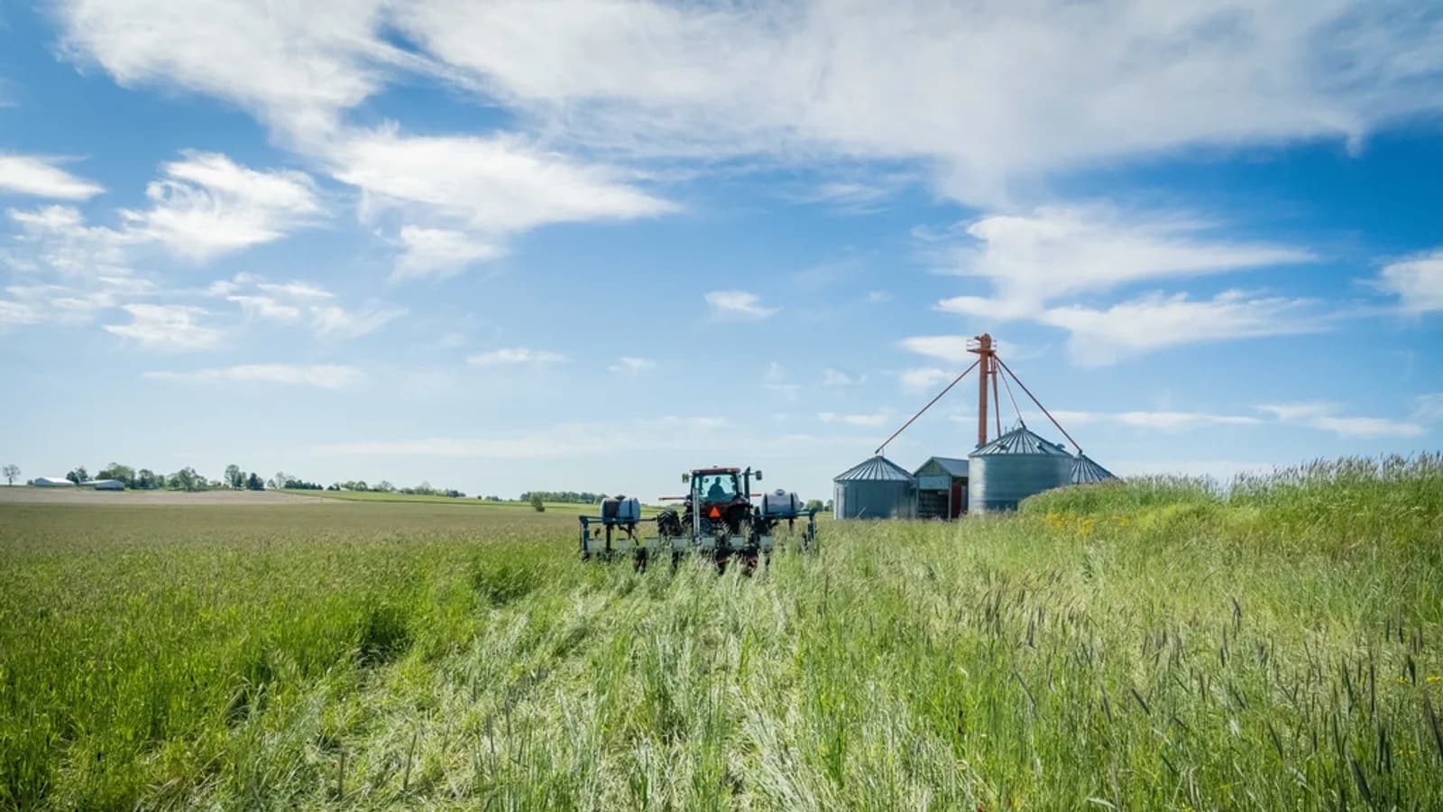 A tractor with a sprayer working a green field, with grain bins and a farmyard in the distance under a blue sky