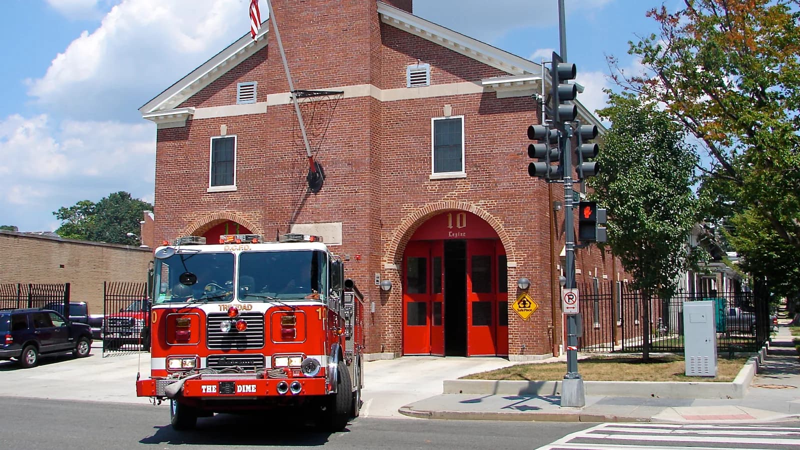 A red fire engine pulling out of a brick fire station's open bay door on a sunny street