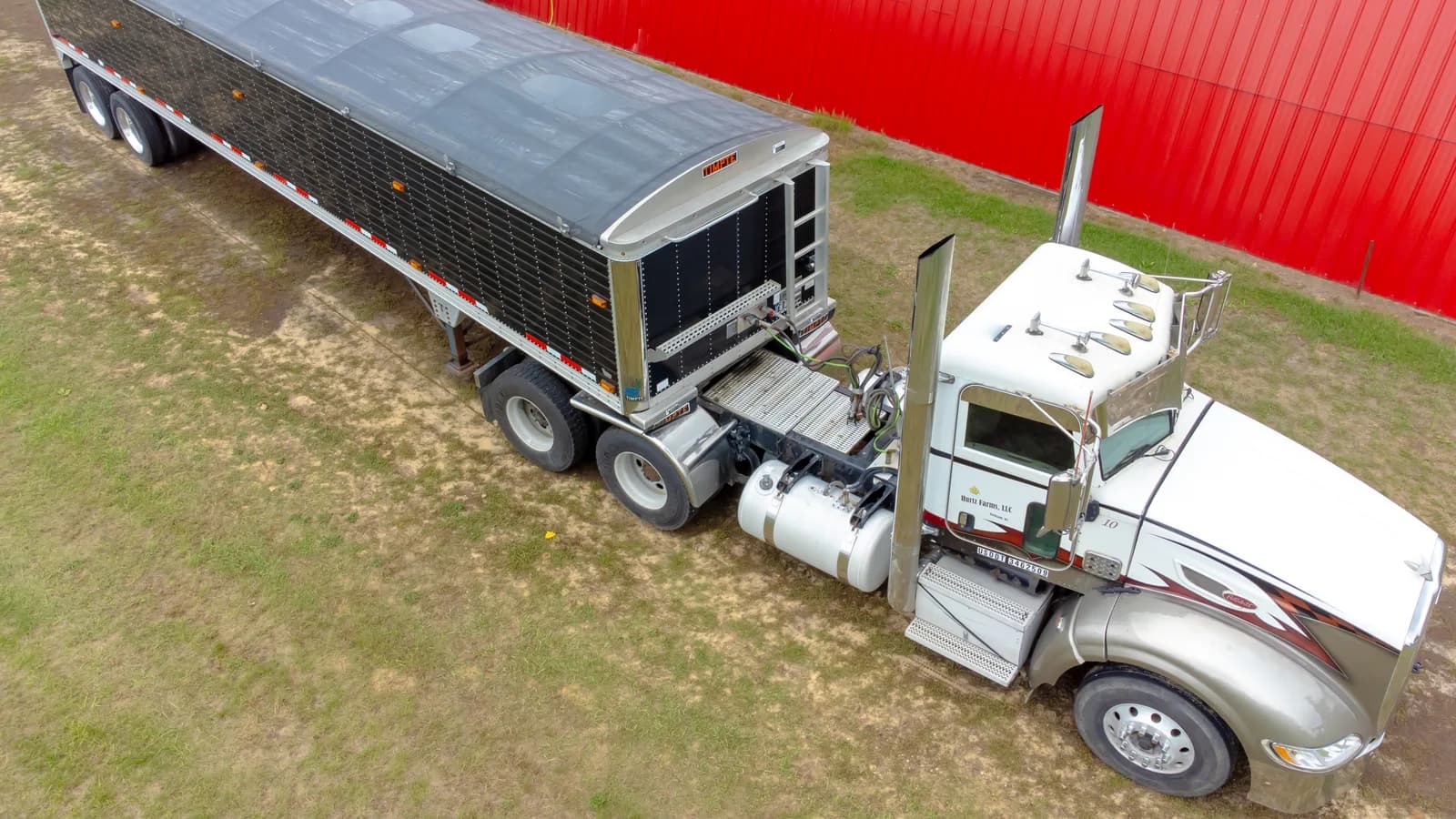 An aerial view of a semi tractor pulling a grain hopper trailer beside a red barn