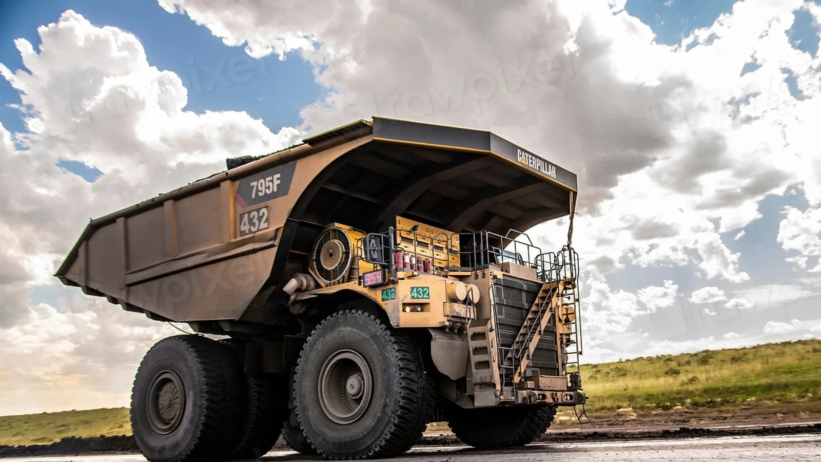 A large yellow mine haul truck on a dirt road under a cloudy sky, with a grassy slope behind it