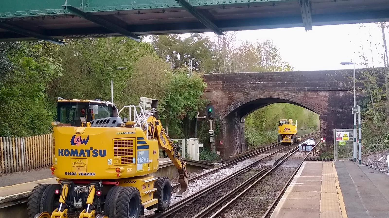 A yellow road-rail excavator working on a track beside a station platform