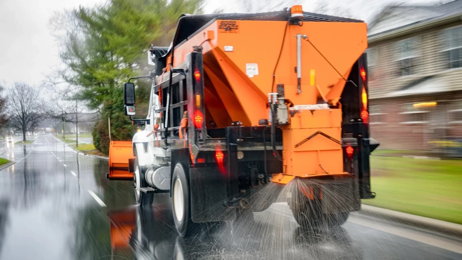 An orange plow truck spreading salt on a wet street, seen from behind as it drives past bare trees